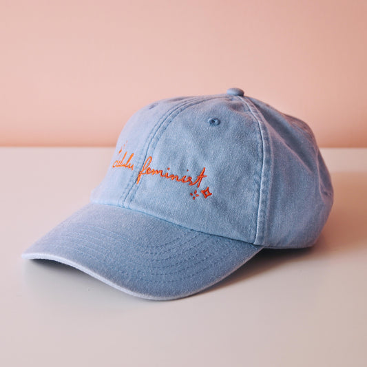 seitliche Ansicht einer hellblauen Cap mit dem aufgestickten Schriftzug "Delulu Feminist" in orange