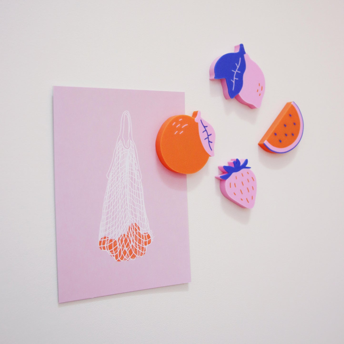 colorful 3D-printed magnets in the shape of fruit holding up a pink postcard