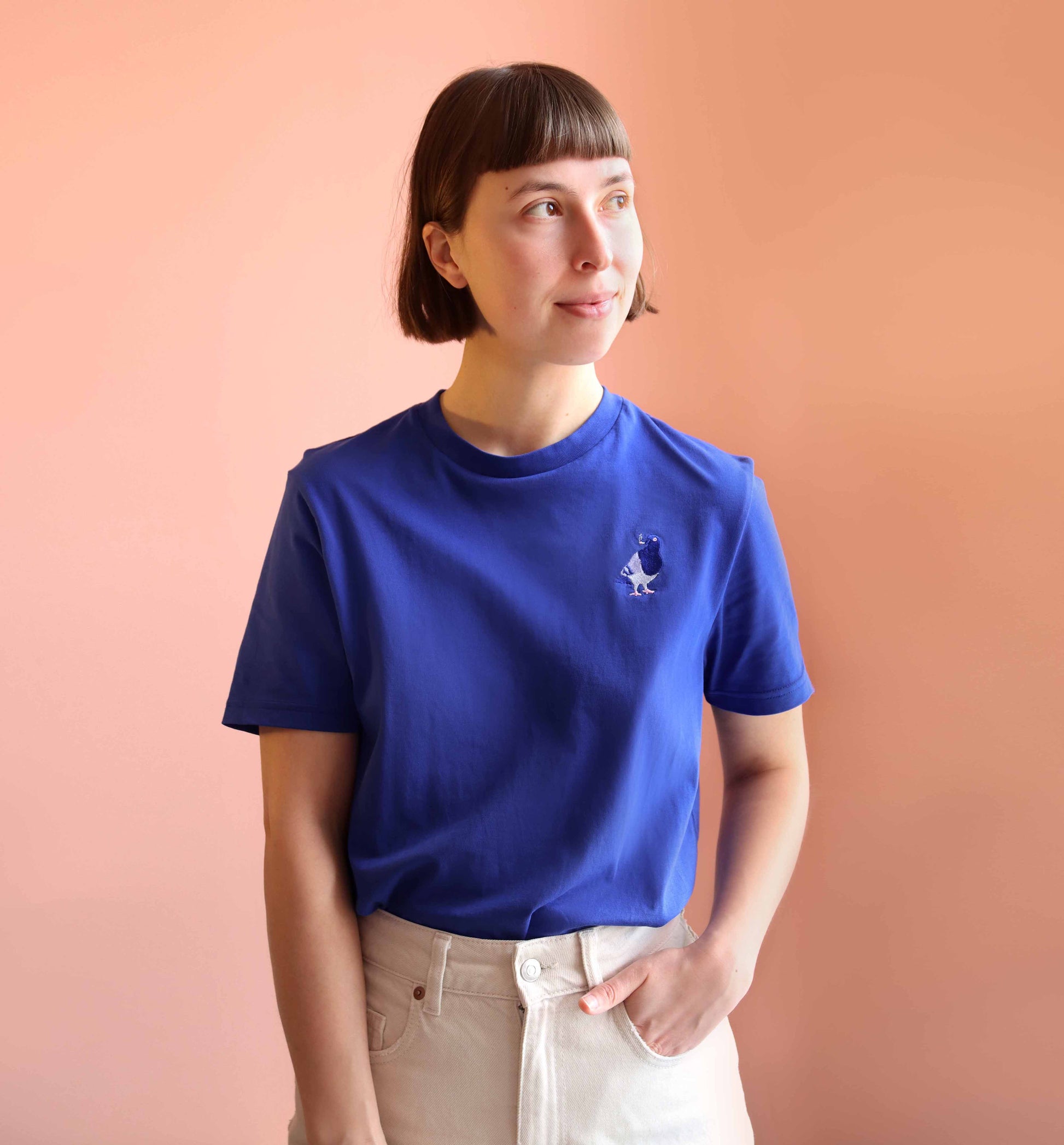 Model trägt blaues Unisex T-Shirt mit einer aufgestickten Taube auf der Brust, die eine Zigarette raucht