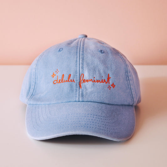 frontale Ansicht einer hellblauen Cap mit dem aufgestickten Schriftzug "Delulu Feminist" in orange 