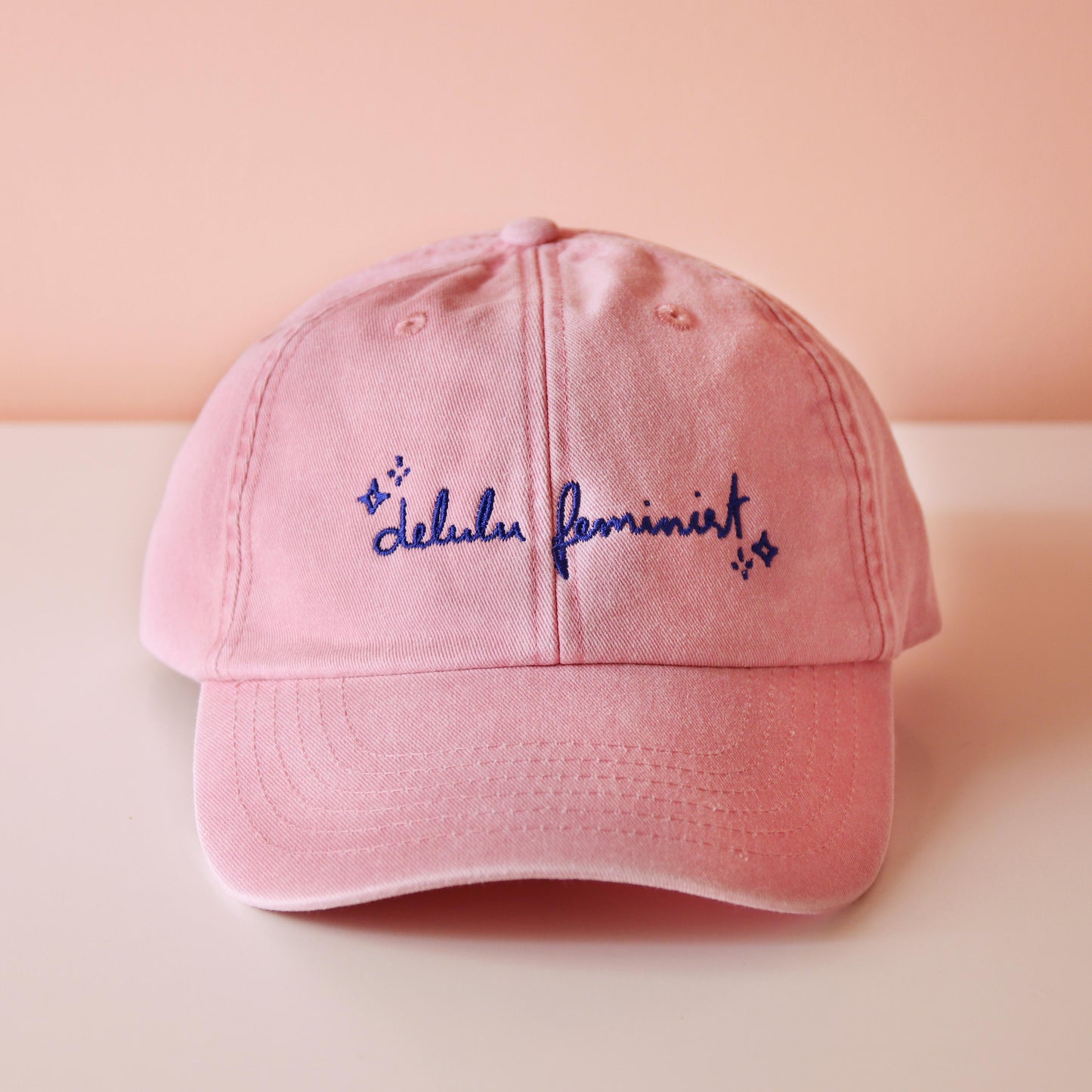 frontale Ansicht einer rosa Cap mit dem aufgestickten Schriftzug "Delulu Feminist" in blau 