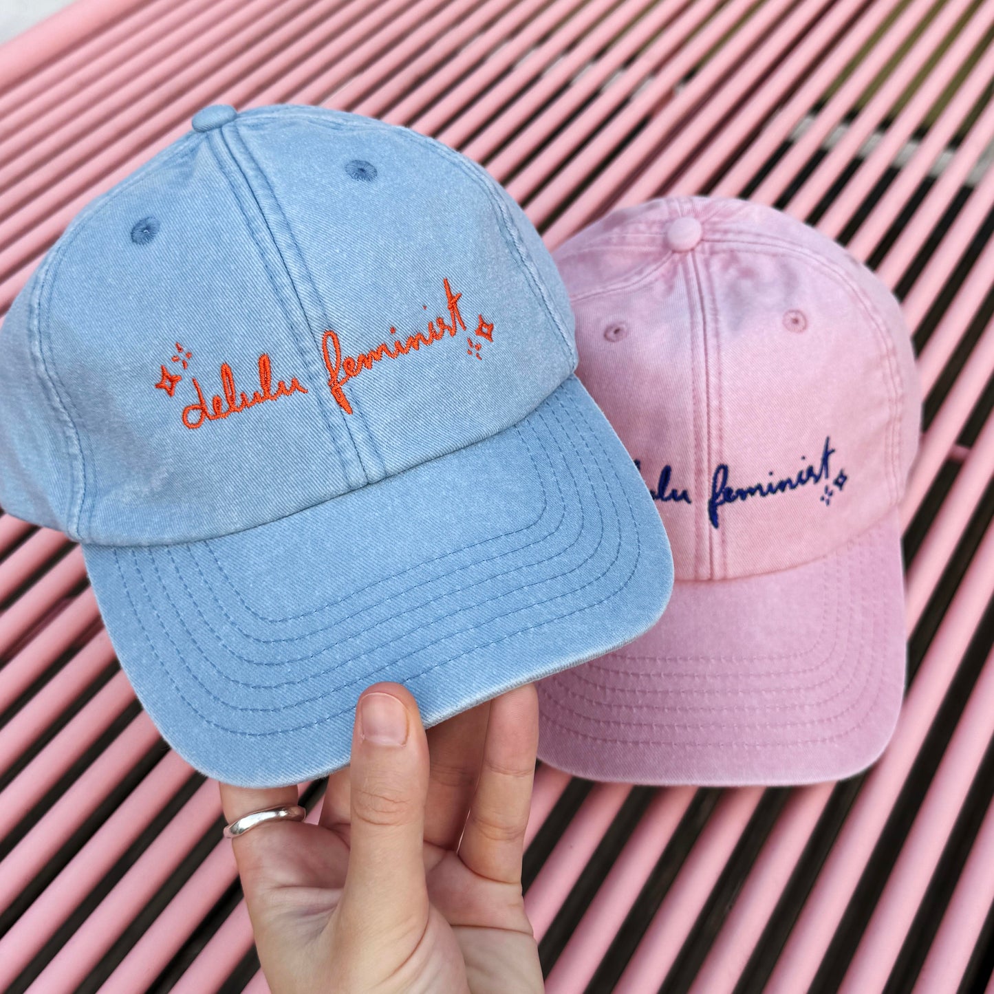 Caps in hellblau und rosa mit dem aufgestickten Text "Delulu Feminist"