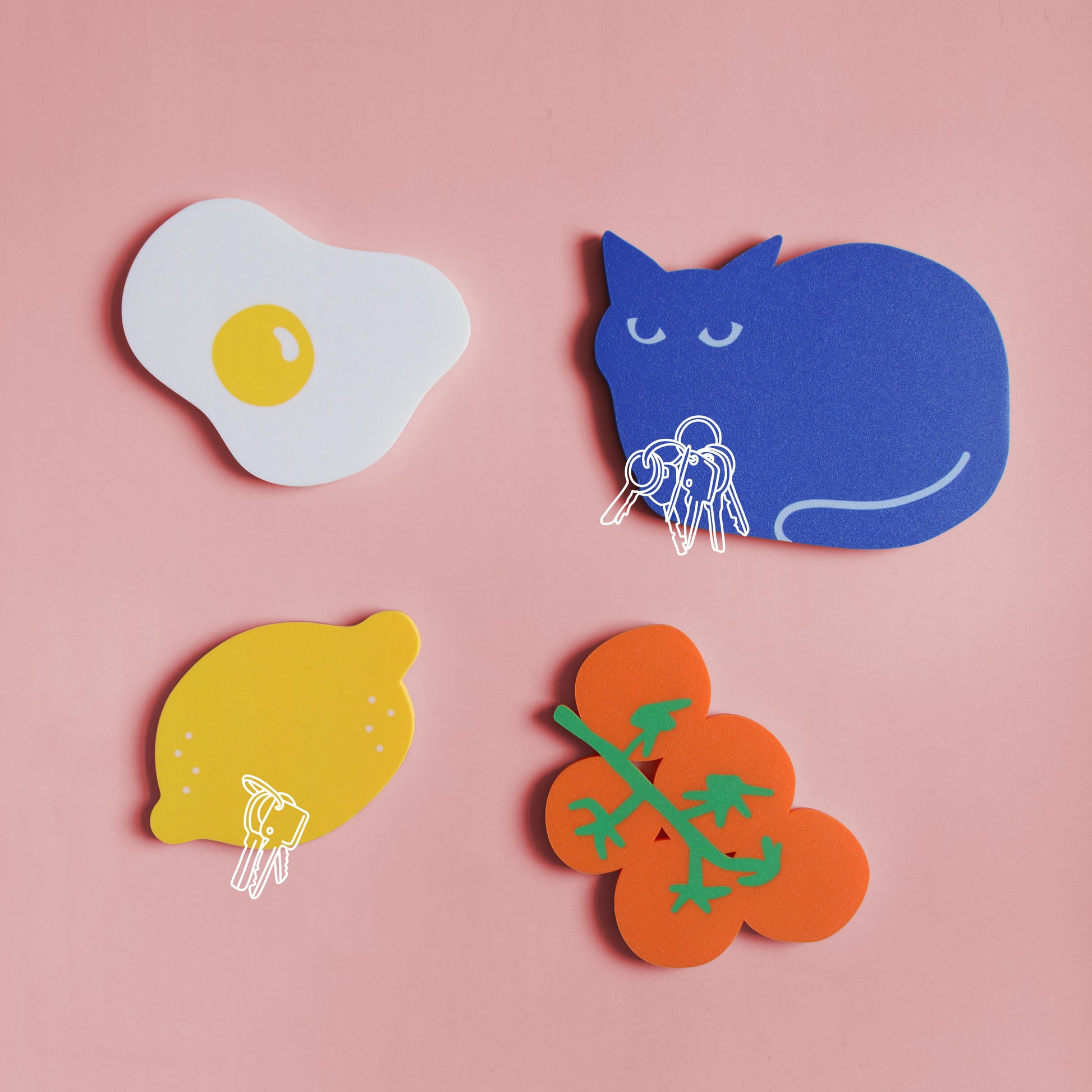 overview of different colorful designs of magnetic key holder such as a fried egg, a blue cat, a lemon and a bunch of tomatoes on a light pink background